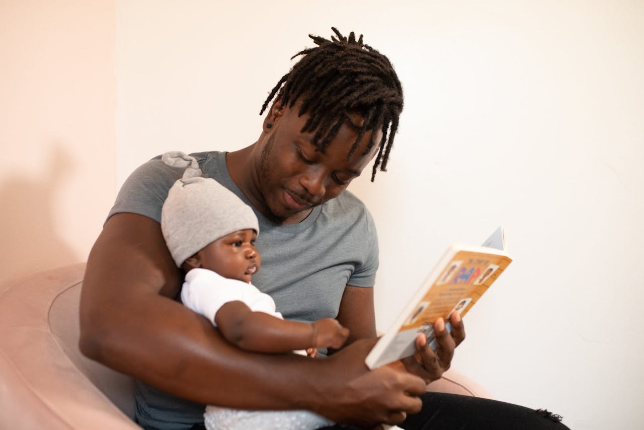 A father with dreadlocks reads a book to his baby, creating a bonding moment.